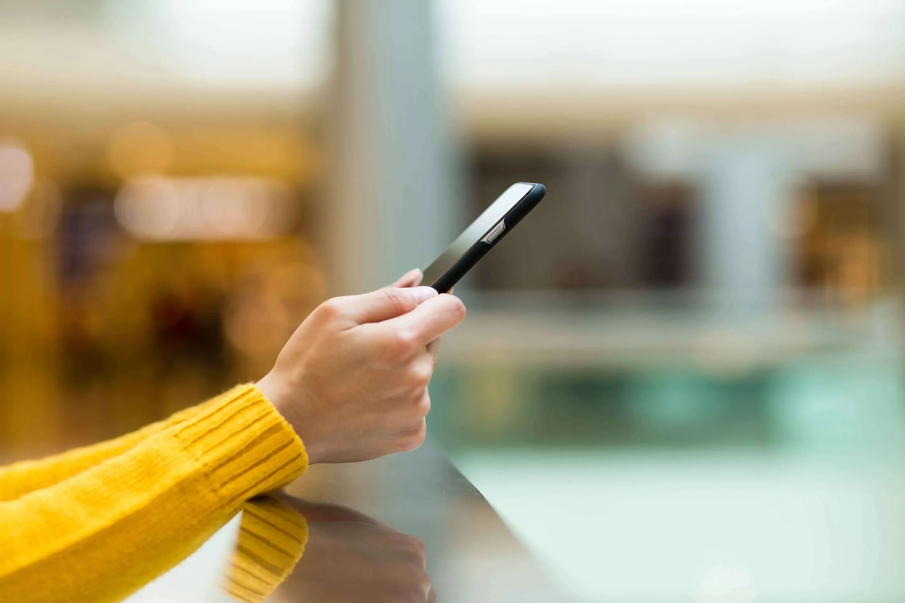A side view of the forearms and hands of a person in a yellow sweater holding a smartphone while leaning against a silver railing. 