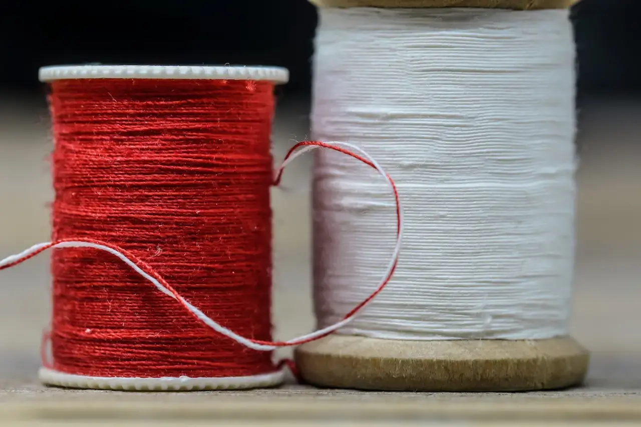 A close-up image of two spools of thread, one red and one white, sitting next to each other on a wood table