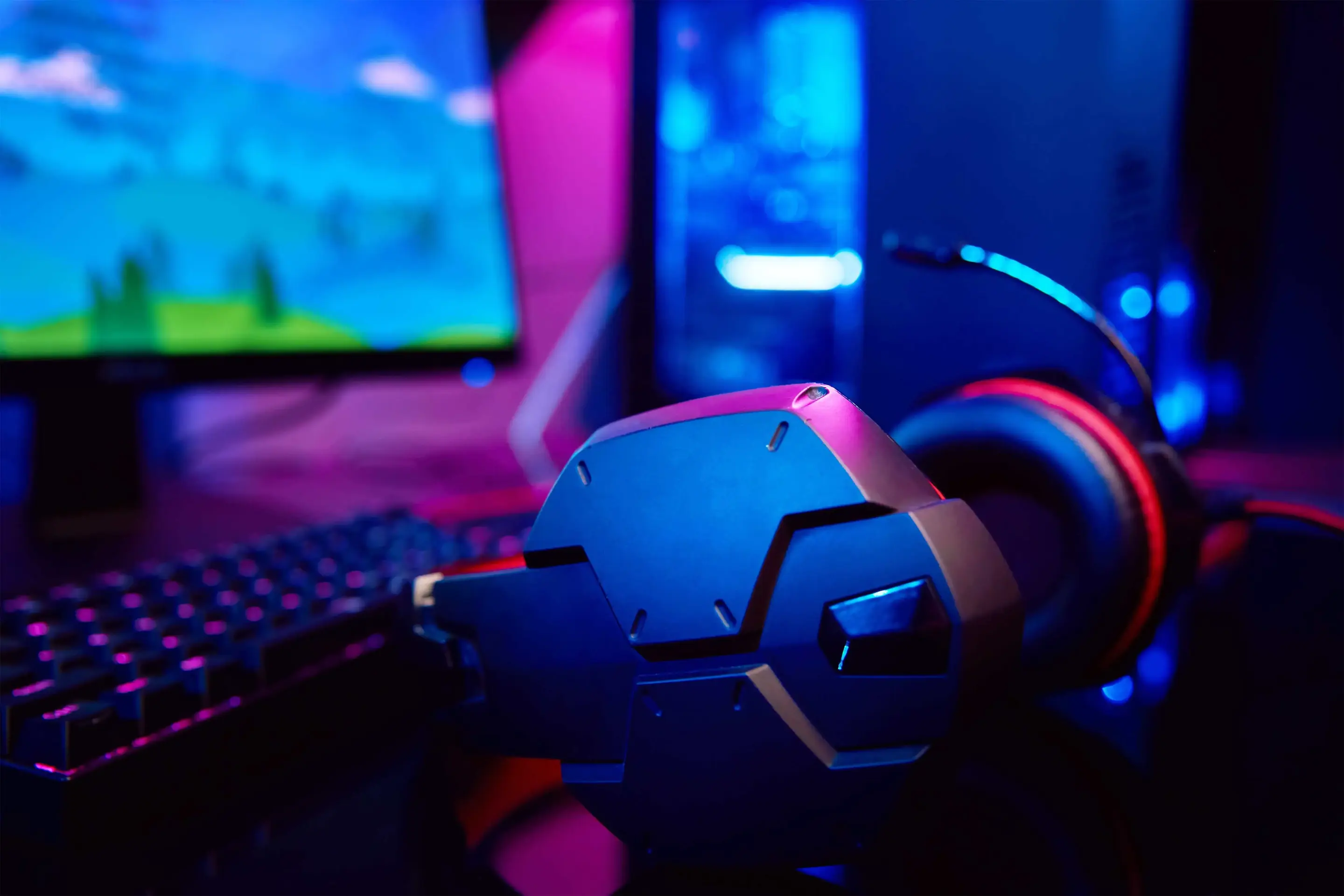 A set of gaming headphones on a desk in front of an LED-lit desktop monitor. 