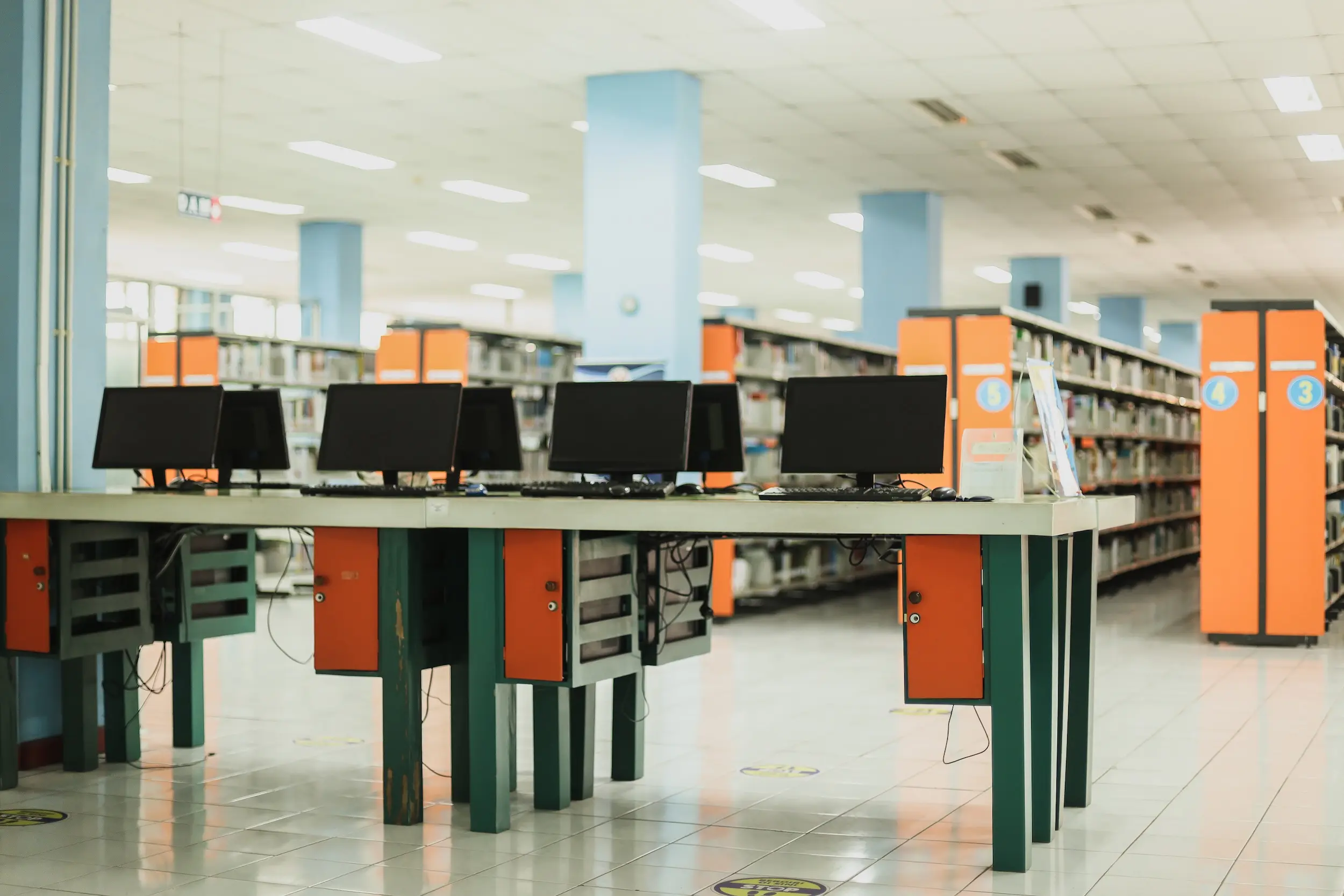 A table of computers in a corner of a library with bookshelves in the background.