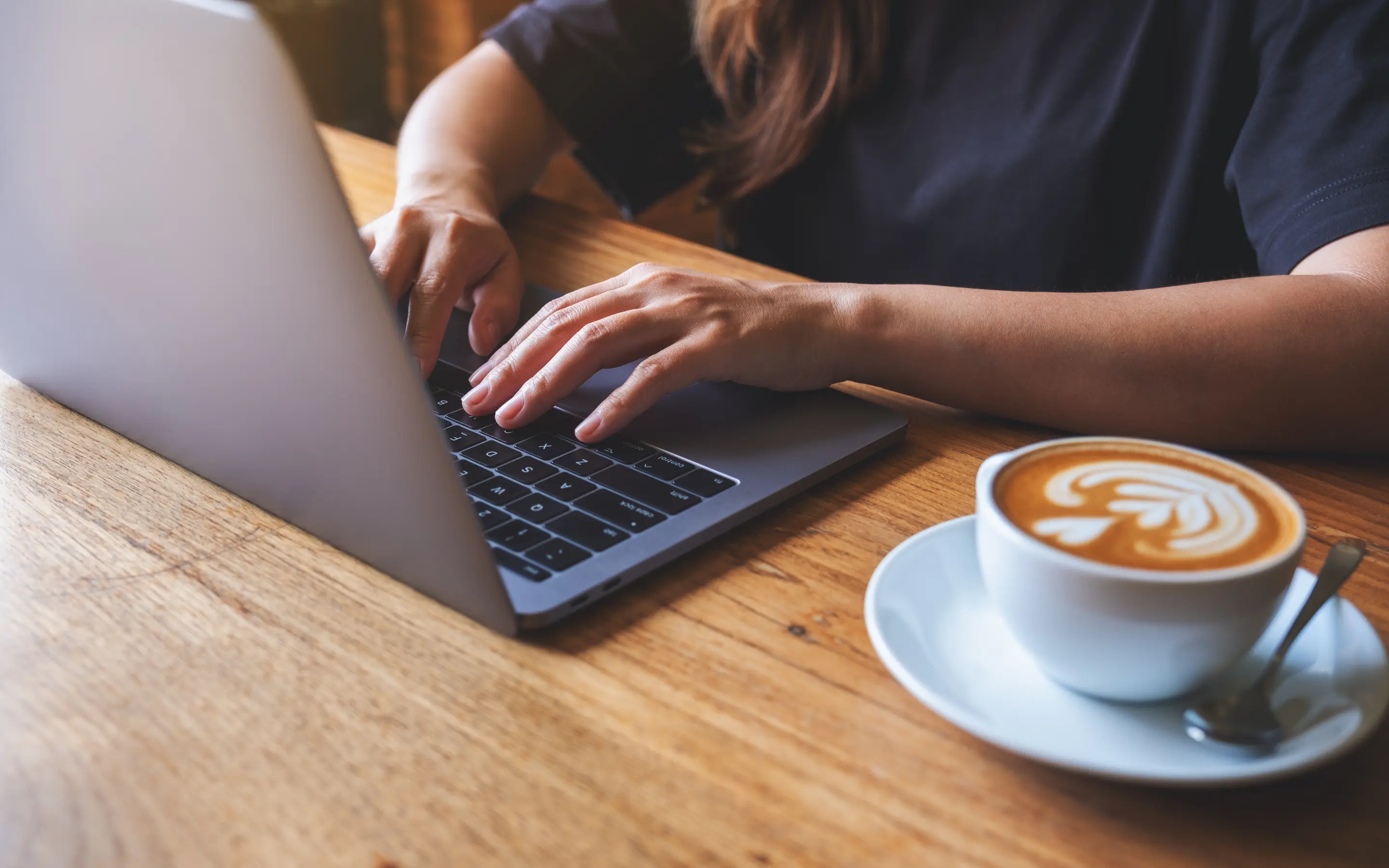 An image of a person's hands on a laptop keyboard with a cup of coffee to their left.