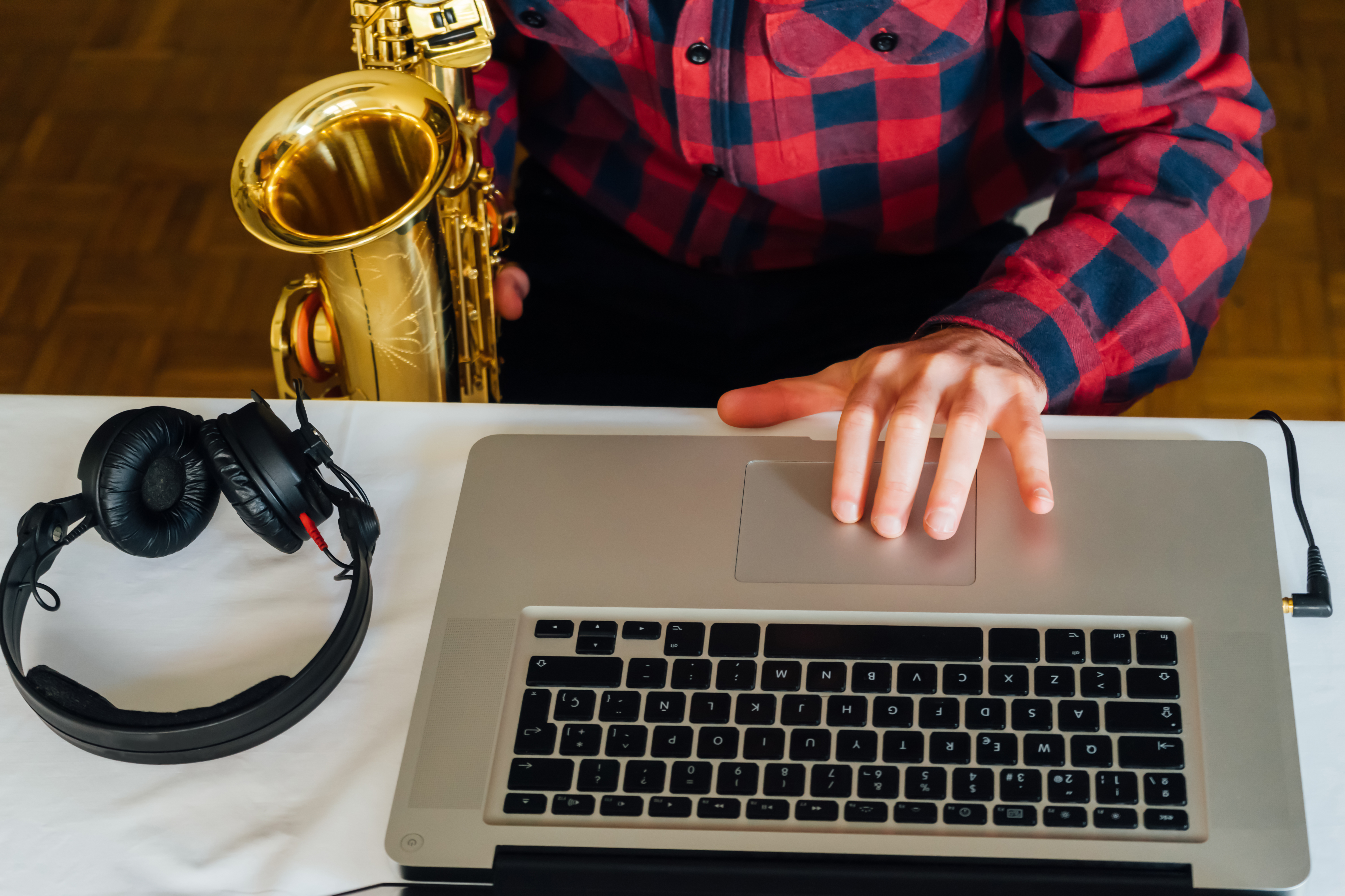 A neck-down image of man in a red flannel shirt holding a saxophone in his right hand, with his left hand on the trackpad of a laptop and headphones on the table next to the laptop. 