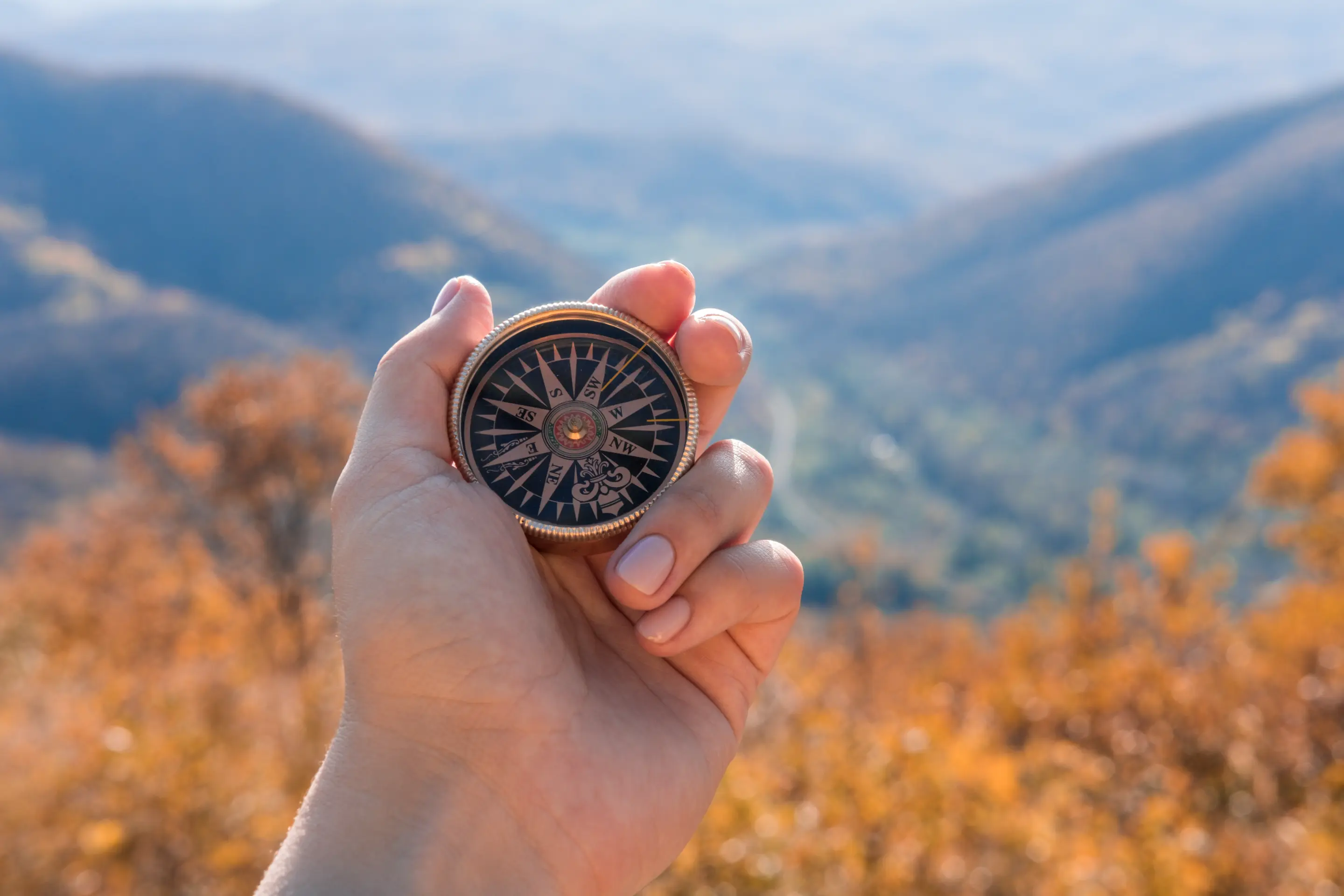 A hand holding a compass in front of a background of fall foliage 